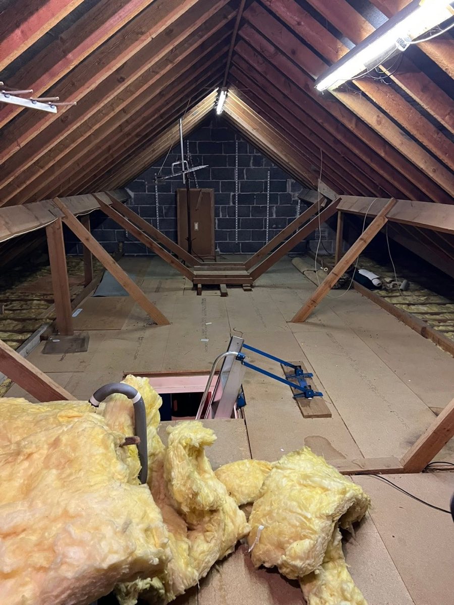 A loft space with exposed roof timbers and rafters, insulation pulled back near the hatch and ladder
