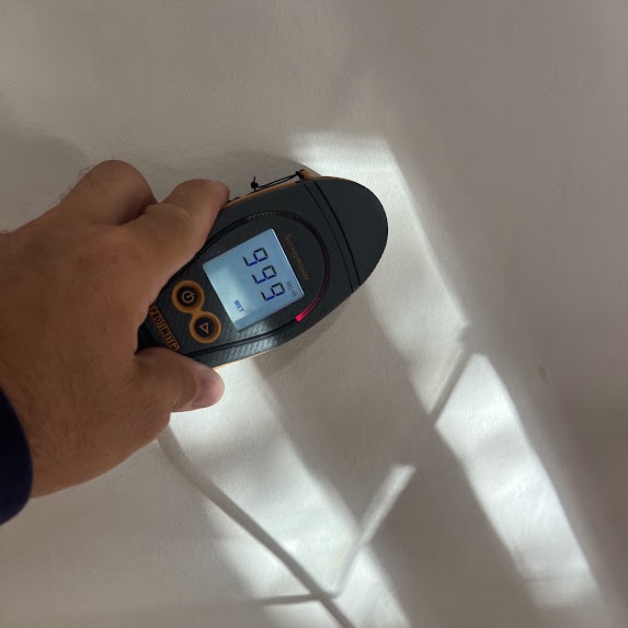 A hand holding a Protimeter moisture meter against a ceiling, the display reading 999, the top of its scale, with the red warning bar lit