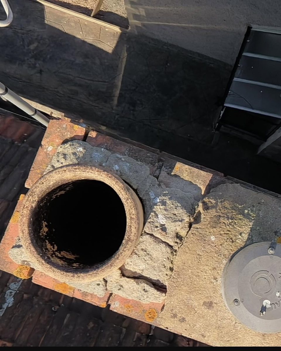 Close view at roof level of a chimney with cracked cement flaunching and a lichen-covered flat roof behind